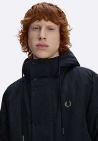 Giacca esterna nera con collo alto, bottoni a pressione, coulisse regolabile e logo ricamato verde sul petto.