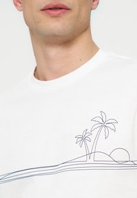 Άνδρας που φοράει λευκό T-shirt με στρογγυλή λαιμόκοψη και μινιμαλιστική μαύρη γραμμή σχεδίασης φοίνικα και ηλιοβασιλέματος στο στήθος.