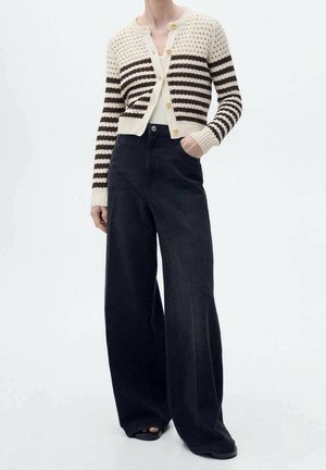 Personne portant un cardigan court rayé crème et marron avec des boutons et un pantalon noir à jambes larges, debout avec une main dans la poche.