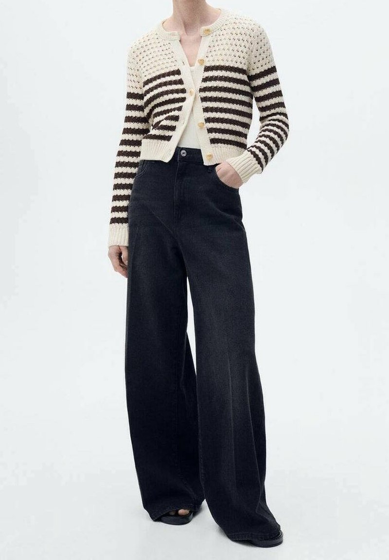 Personne portant un cardigan court rayé crème et marron avec des boutons et un pantalon noir à jambes larges, debout avec une main dans la poche.