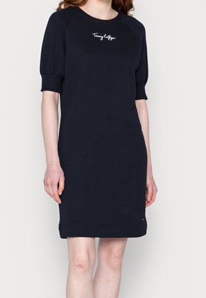 Femme portant une robe bleu marine à manches courtes bouffantes avec "Tommy Hilfiger" brodé sur la poitrine.