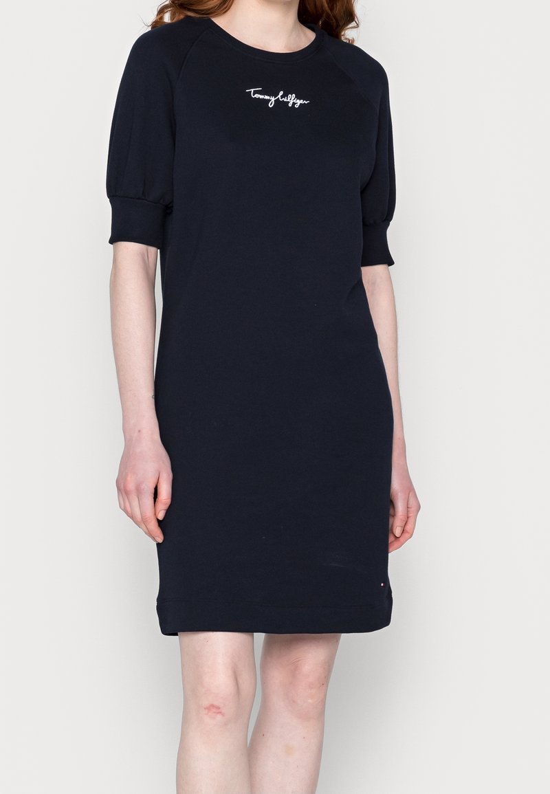 Femme portant une robe bleu marine à manches courtes bouffantes avec "Tommy Hilfiger" brodé sur la poitrine.
