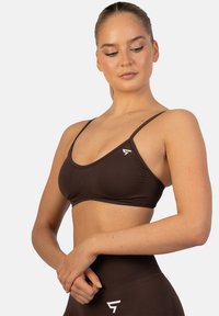 Ruskea urheiluliivi, jossa on ohuet hihnat ja kolmiomainen muotoilu. Vasemmalla puolella on valkoinen logo. Materiaali näyttää sileältä ja istuvalta.