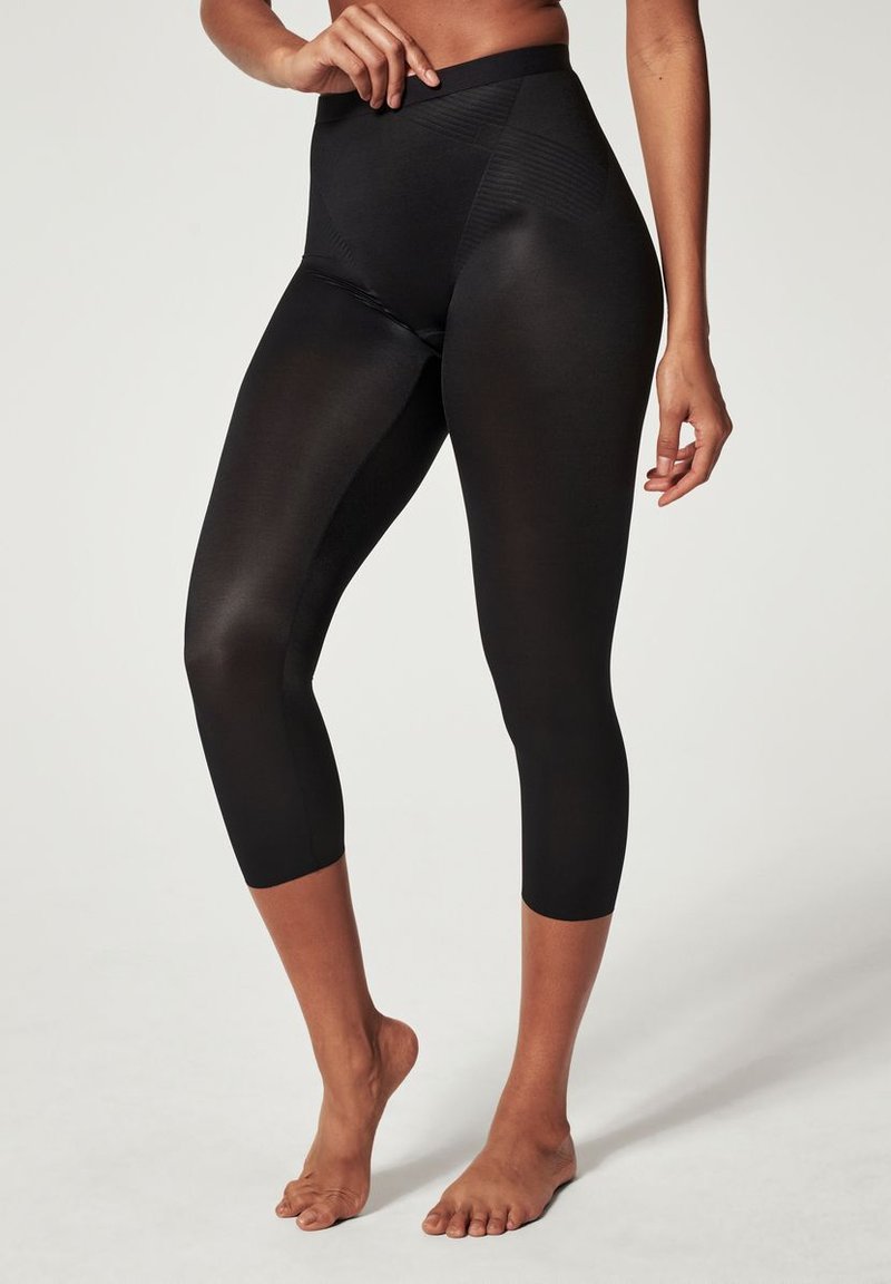 Leggings capri noirs avec une taille haute, fabriqués dans un matériau lisse et extensible, présentant de subtiles accents texturés sur les côtés.