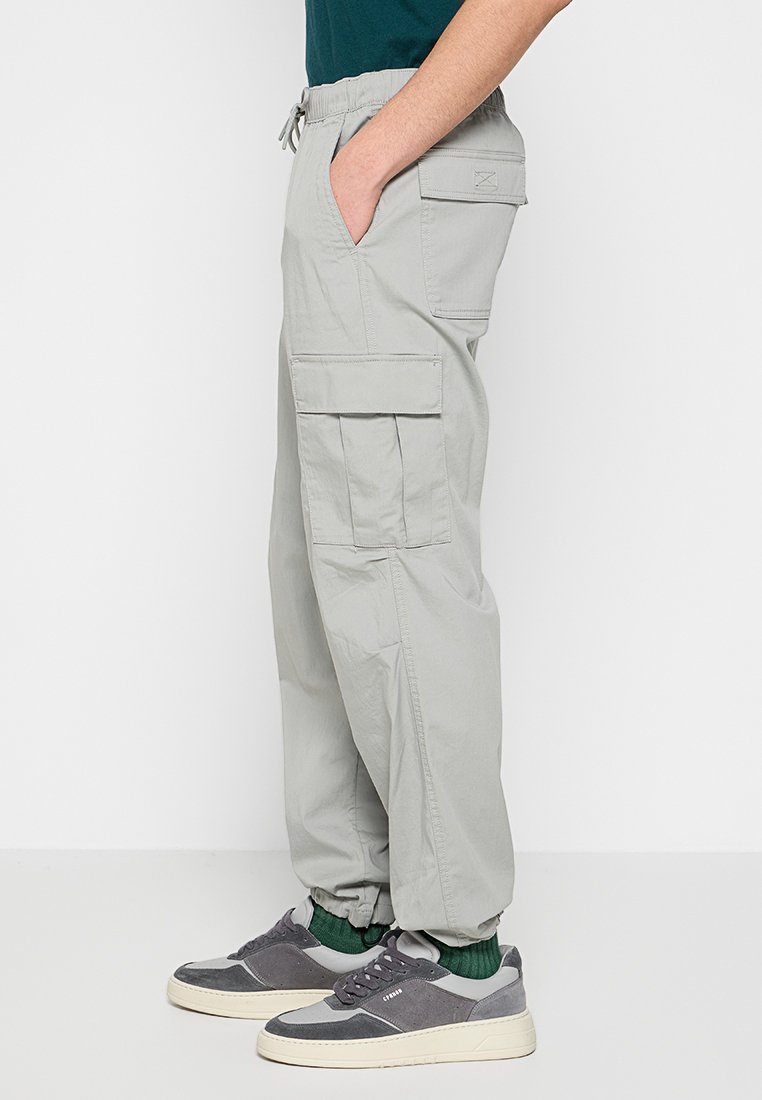 Hellgraue Cargo-Hosen mit elastischem Kordelzugbund, seitlichen Taschen und großzügigem Schnitt. Kombiniert mit grauen Sneakers und grünen Socken.