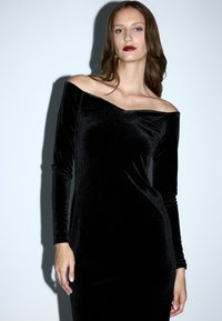 Vestido negro de terciopelo, con escote bardot y mangas largas, silueta ajustada, textura suave y un sutil brillo.