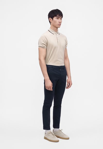 Homme debout portant un polo beige à manches courtes, un pantalon slim foncé, des chaussettes blanches et des baskets beiges sur un fond blanc uni.