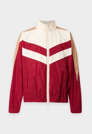Jeune homme portant une veste zippée rouge et beige, un jean noir et des baskets rouges, debout devant un fond blanc uni.