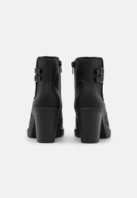 Botas de tobillo de cuero negro con tacones de bloque, que presentan dos hebillas decorativas laterales. La textura es suave con sutiles detalles de costura.