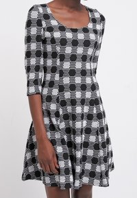 Czarna i biała sukienka w kropki z dopasowaną górą i rozkloszowanym dołem, z rękawami o długości trzech czwartych i dekoltem w serek.