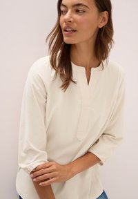Blusa beige chiaro con scollo a barchetta e spacco frontale. Presenta maniche a tre quarti con polsini elastici e un tessuto morbido e testurizzato.
