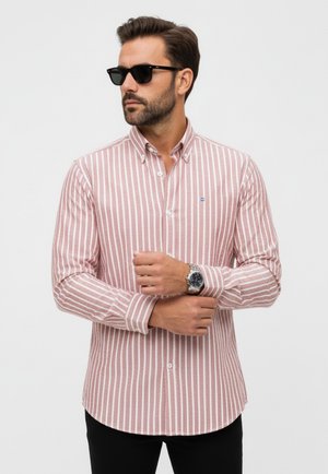 Camisa de botones a rayas rojas y blancas con cuello, de manga larga y un pequeño logo azul. Llevada con pantalones oscuros y un reloj de pulsera.