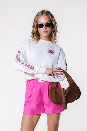 Hvid langærmet skjorte med pinkt grafisk motiv og tekst, kombineret med teksturerede pink shorts og en brun ruskindstaske med kvast.