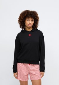 Fekete kapucnis pulóver vörös logós emblémával, rózsaszín, szívmintás rövidnadrággal párosítva, mutatva a laza szabást és a puha textúrát.