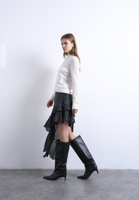 Γυναίκα που περπατάει, φορώντας λευκό τοπ με μακριά μανίκια, μαύρη ασύμμετρη φούστα με πουά και μαύρες δερμάτινες μπότες με τακούνι μέχρι το γόνατο.