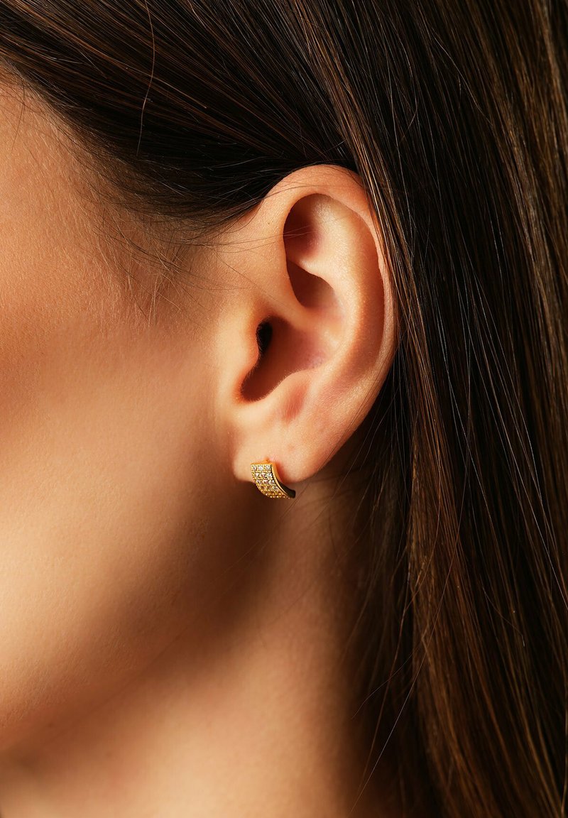 Nahaufnahme eines Frauenohres mit glattem, dunklem Haar, das ein kleines goldfarbenes Ohrstecker mit eingearbeiteten klaren Edelsteinen trägt.