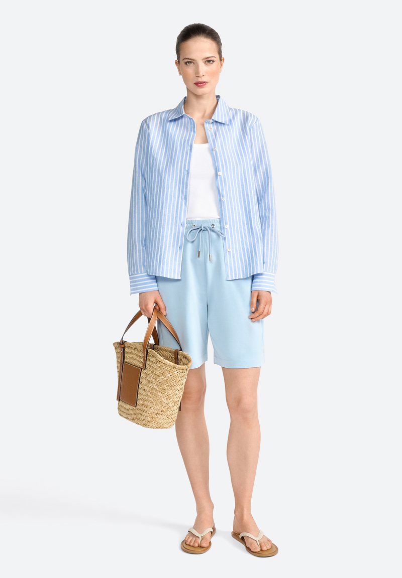 Chemise à rayures bleu clair sur un débardeur blanc, associée à un short bleu doux. Un sac en paille tressé avec des accents marron complète le look.
