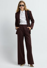Bruin gestreept kostuum met wijde pijpen en een op maat gemaakt jasje, gecombineerd met een witte blouse en spitse gouden hakken. Model draagt een zonnebril.