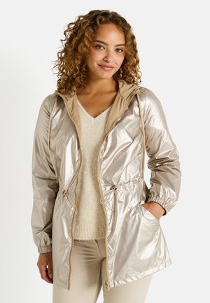 Femme aux cheveux bouclés portant une veste à capuche zippée beige brillante, un pull crème et un pantalon clair, se tenant avec une main dans la poche.