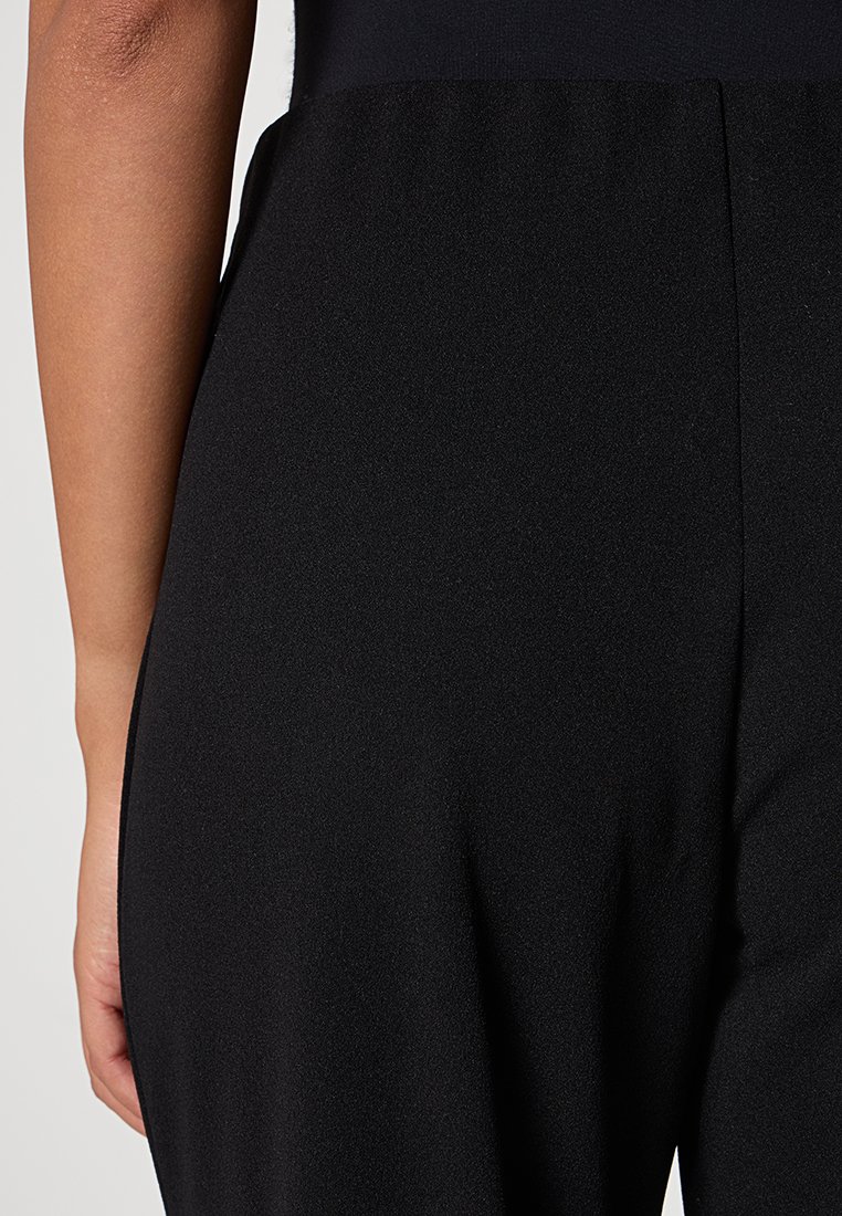 Gros plan sur le bas du torse et le bras d'une personne portant un pantalon noir ajusté devant un fond neutre.