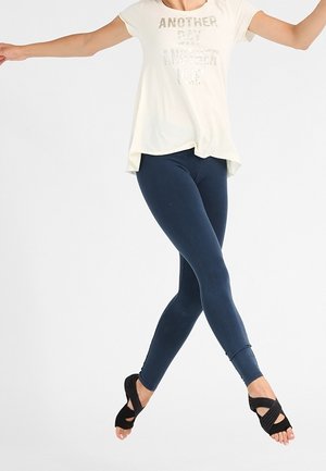 Kvinde i hvid skjorte og marineblå leggings springer midt i luften med armene udstrakte, iført sorte åbne danseko på en hvid baggrund.