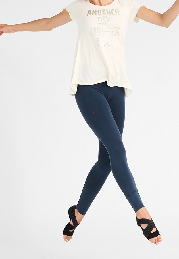Femme en chemise blanche et leggings bleu marine sautant en l'air, les bras étendus, portant des chaussures de danse noires à bout ouvert sur fond blanc.