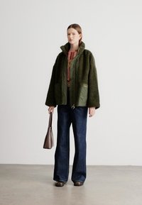 Roheline, pehme paksudega jope kõrge kraega ja kahe eesmise taskuga, kantud punase mustriga särgi peal, kombineeritud tumedate laiade teksade ja pruuni kotiga.