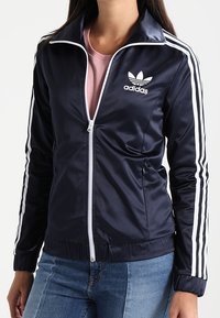 Giacca con zip blu navy realizzata in tessuto liscio e lucido con dettagli a tre strisce bianche sulle maniche e logo ricamato sul petto.