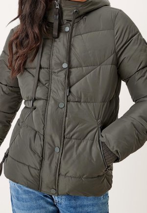 Kvinde iført en olivengrøn quiltet jakke med trykknapper, lynlåslommer og hætte, kombineret med blå jeans.