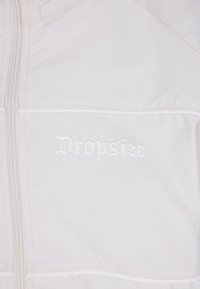 Giacca bianca con chiusura a zip realizzata in tessuto leggero, dotata di una tasca frontale e un logo ricamato "Dropsize" in un colore a contrasto.