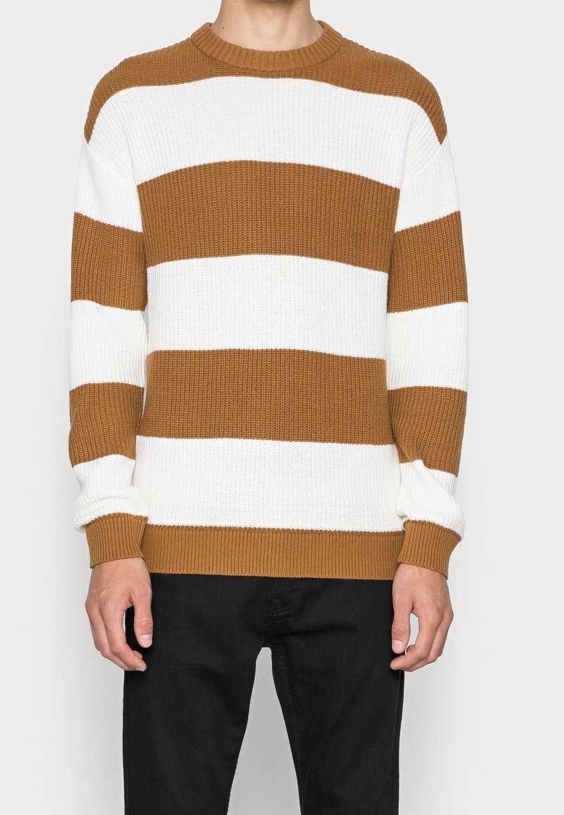 Personne portant un pull en tricot à larges rayures marron et blanc et un pantalon noir, les mains détendues le long du corps.