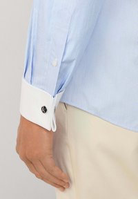 Braccio maschile che indossa una camicia azzurra a righe sottili con polsini bianchi e gemelli argentati, accanto a pantaloni beige.