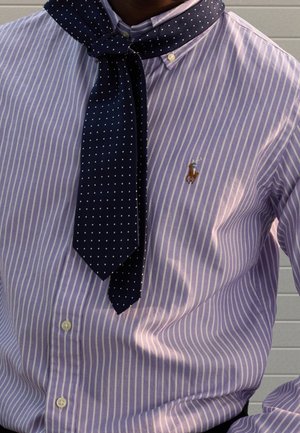 Mężczyzna w koszuli w paski w kolorach lawendy i bieli, z granatowym szalikiem w groszki zawiązanym wokół kołnierza.