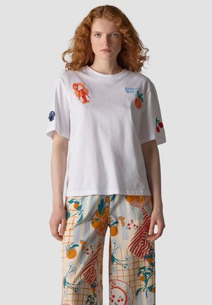 Woman with red curly hair wearing a white t-shirt with fruit and seafood embroidery and colorful patterned wide-leg pants.
