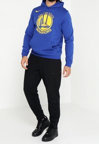 Sudadera azul con el logo de los Golden State Warriors y un bolsillo tipo canguro en la parte frontal; combinada con pantalones jogger negros y zapatillas deportivas negras.