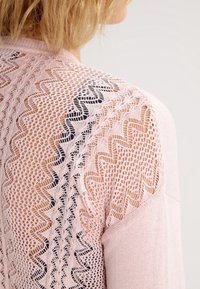 Rosa stickad tröja med ett zick-zack mönster av spets på baksidan, med kontrasterande svarta detaljer och en mjuk textur.