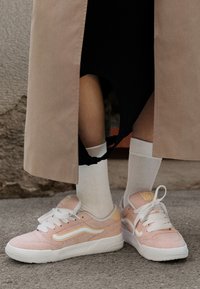 Rosa Wildledersneaker mit weißen Akzenten und Schnürsenkeln, ausgestattet mit einem beigen Rückenpanel. Getragen mit weißen Socken und einem langen beigen Mantel.