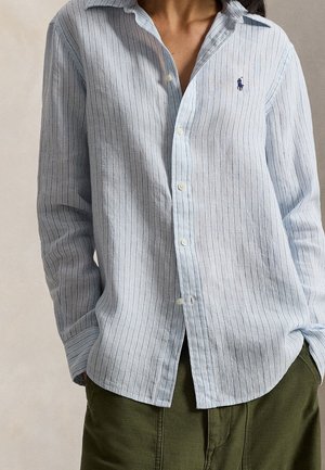 Chemise bleu clair à manches longues avec fines rayures verticales portée rentrée dans un pantalon vert olive, mains dans les poches, col et boutons visibles.