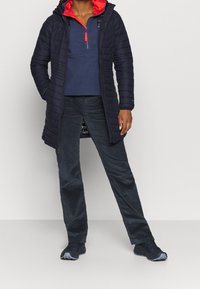 Marinblå lång pufferjacka med kviltad design, rödfodrad huva, marinblå fleece-tröja, mörka byxor och svarta sportskor.