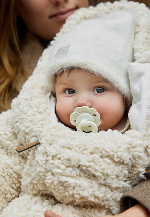 Beige fluffy outerwear envelops a child with a white knitted hat. A flower-shaped pacifier rests in the child's mouth, revealing blue eyes.