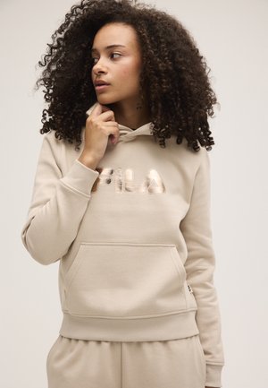 Mujer con el pelo rizado que lleva una sudadera con capucha beige de FILA y pantalones a juego, mirando hacia un lado con la mano cerca del mentón.