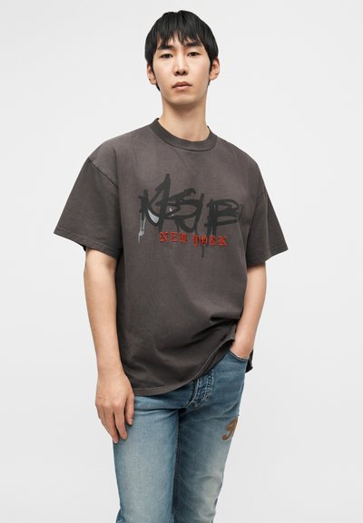 Μεγάλο γκρι σκούρο μπλουζάκι με γραφικό κείμενο σε μαύρο και κόκκινο, συνδυασμένο με τζιν παντελόνι ανοιχτό μπλε. Το ύφασμα φαίνεται λείο και ελαφρύ.