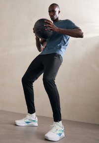 Herreidrettsantrekk: blå kortermet skjorte, svarte slim-fit bukser og hvite joggesko med grønne og blå detaljer. Holder en svart ball.