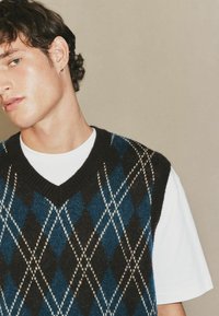 Blaue und schwarze Argyle-Strickweste mit weißen Akzenten, featuring einen V-Ausschnitt und ärmelloses Design, über einem weißen T-Shirt getragen.
