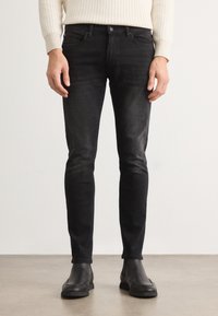 Jeans de mezclilla negra ajustados con acentos desgastados, presentan un diseño tradicional de cinco bolsillos y costuras reforzadas. Combinados con botas negras hasta el tobillo.