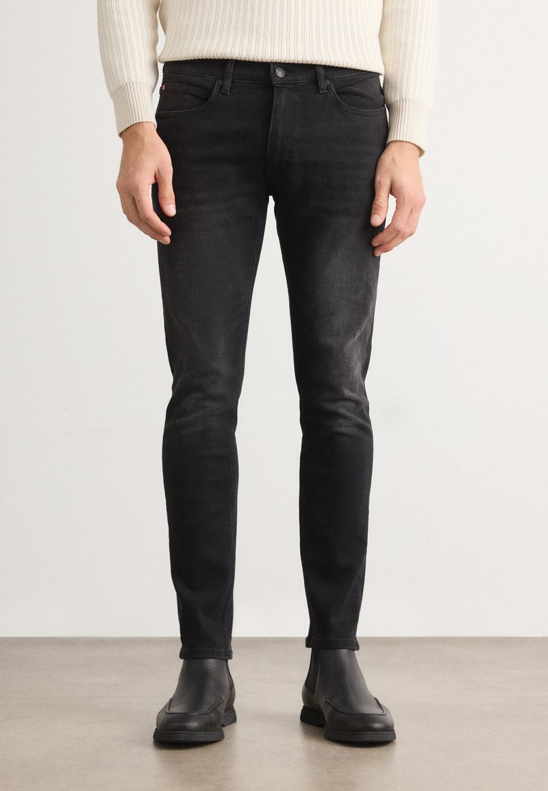 Jeans de mezclilla negra ajustados con acentos desgastados, presentan un diseño tradicional de cinco bolsillos y costuras reforzadas. Combinados con botas negras hasta el tobillo.