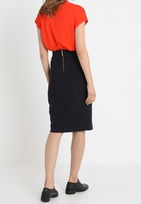 Červený top s krátkým rukávem v kombinaci s černou tužkovou sukní se zipem na zadní straně. Model v černých botách, pohled zezadu.