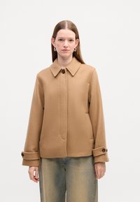Brun struktureret jakke med krave, knaplukning og knapmanchetter, parret med falmede blå jeans. Glat stof og struktureret design.