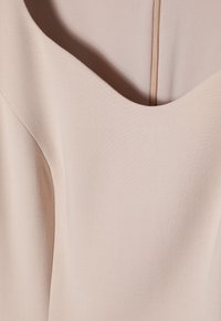 Beige klänning med djup rundad halsringning, tillverkad av ett mjukt tyg; har subtil textur och en dold dragkedja på baksidan.