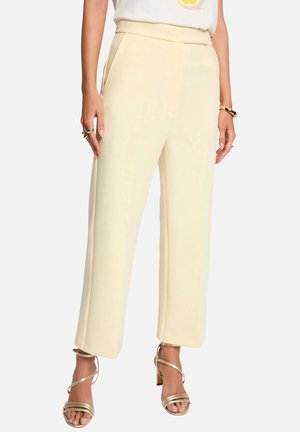 Femme portant un pantalon crème taille haute à jambes larges avec poches latérales, associé à des sandales à talons hauts dorées à brides et des bijoux en or.